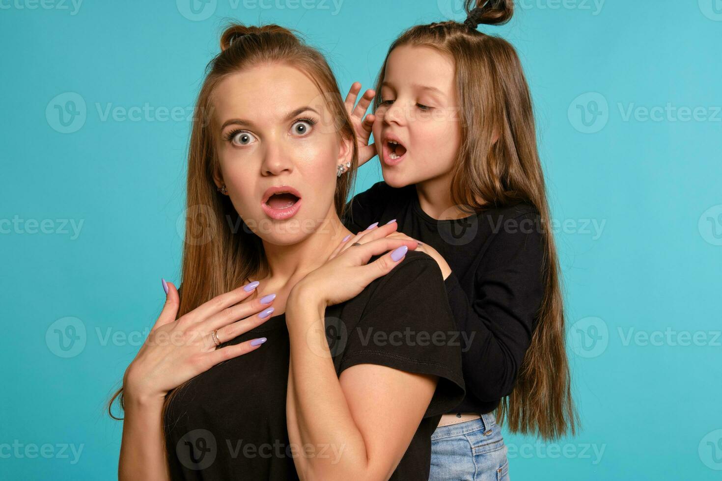 mamá y hija con un gracioso peinados, vestido en negro camisas y azul mezclilla pantalones son posando en contra un azul estudio antecedentes. de cerca disparo. foto