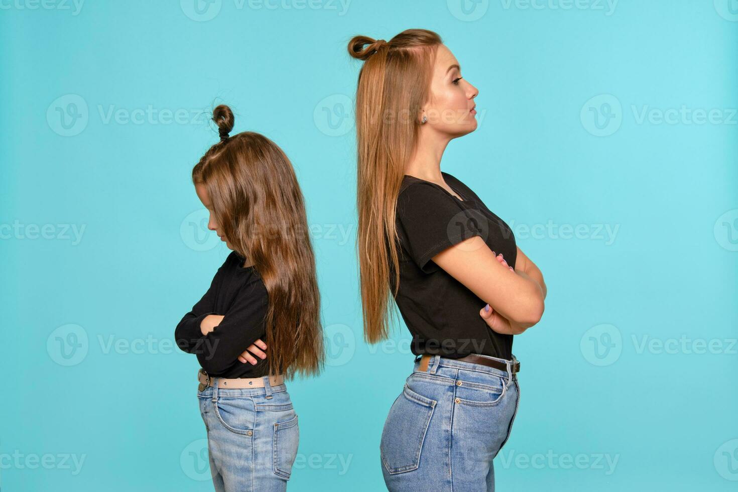 mamá y hija con un gracioso peinados, vestido en negro camisas y azul mezclilla pantalones son posando en contra un azul estudio antecedentes. de cerca disparo. foto