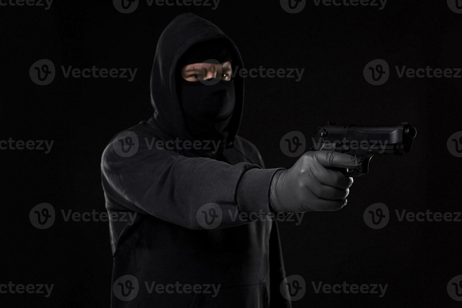 The robber in a mask with a gun pointed to the side on a black background photo