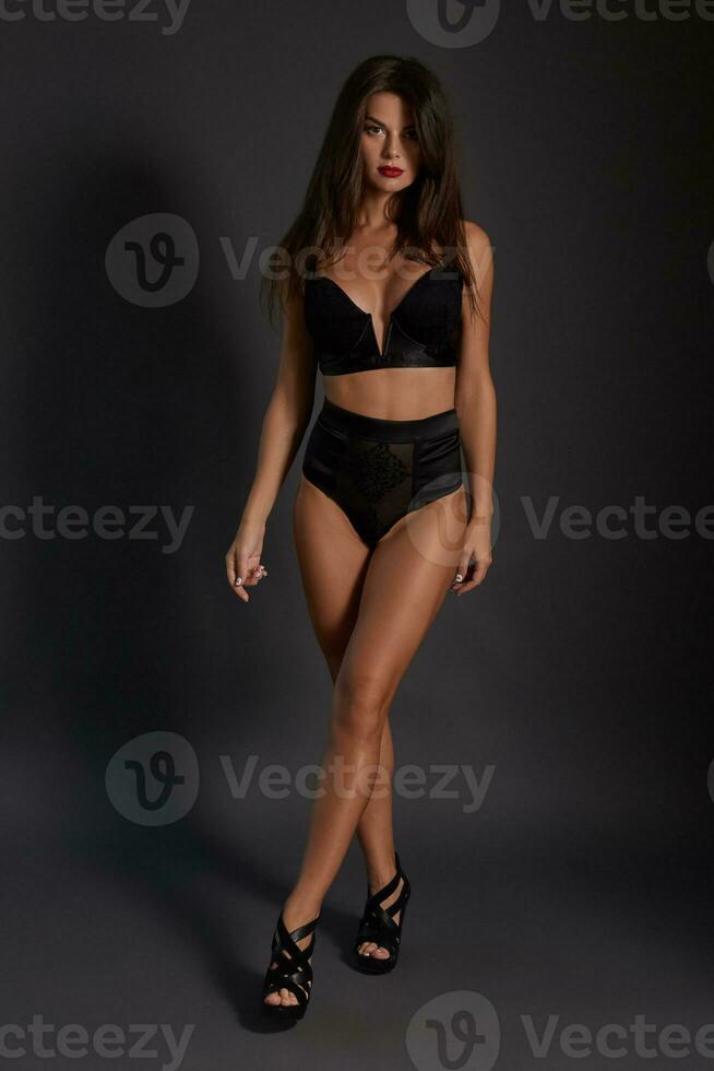 Young sexy slim tanned woman in black underwear posing against dark grey background. photo