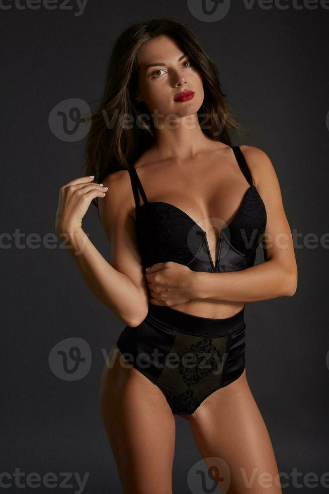 Stunning caucasian female model with dark hair and red lips in black underwear posing on dark grey background. photo