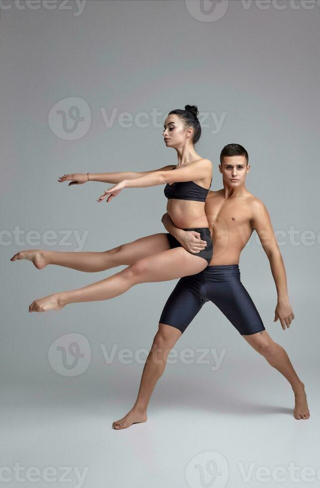 The couple of a young modern ballet dancers in black suits are posing over a gray studio background. photo