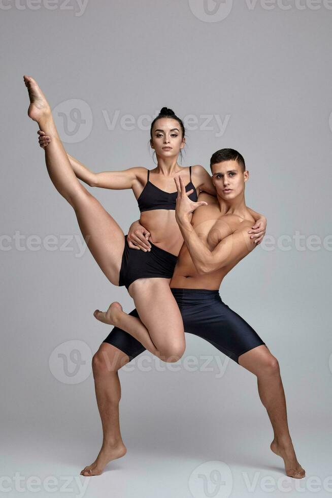 The couple of a young modern ballet dancers in black suits are posing over a gray studio background. photo