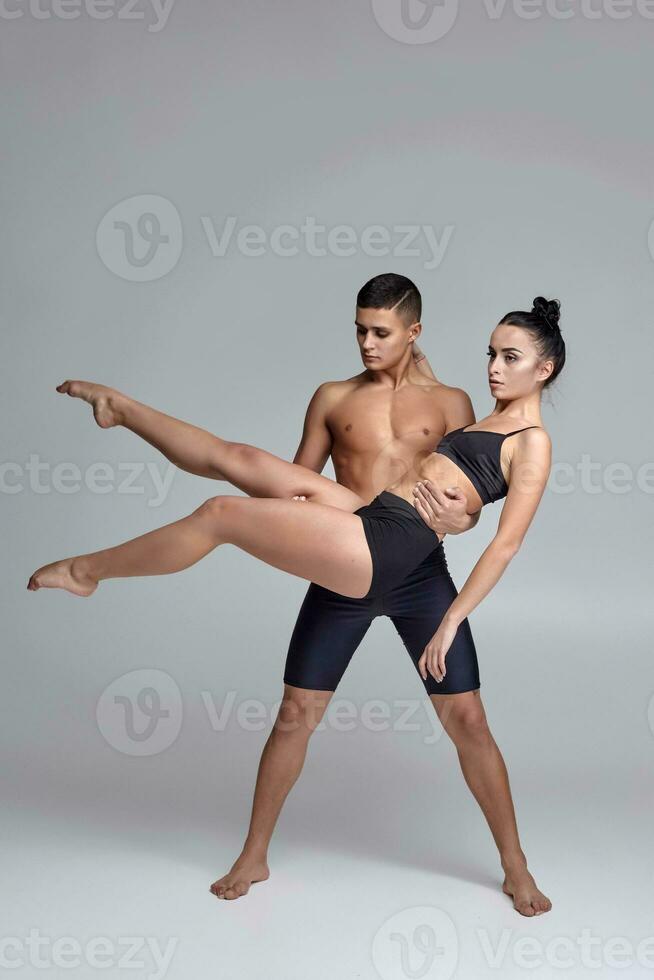 The couple of a young modern ballet dancers in black suits are posing over a gray studio background. photo