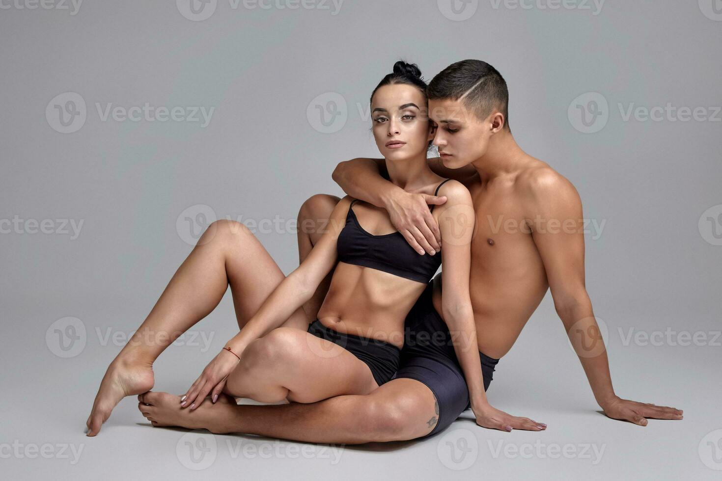 The couple of a young modern ballet dancers in black suits are posing over a gray studio background. photo