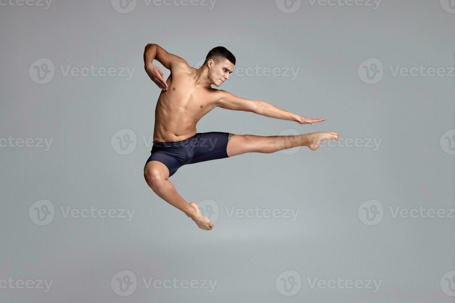 Photo of a handsome man ballet dancer, dressed in a black shorts, making a dance element against a gray background in studio.