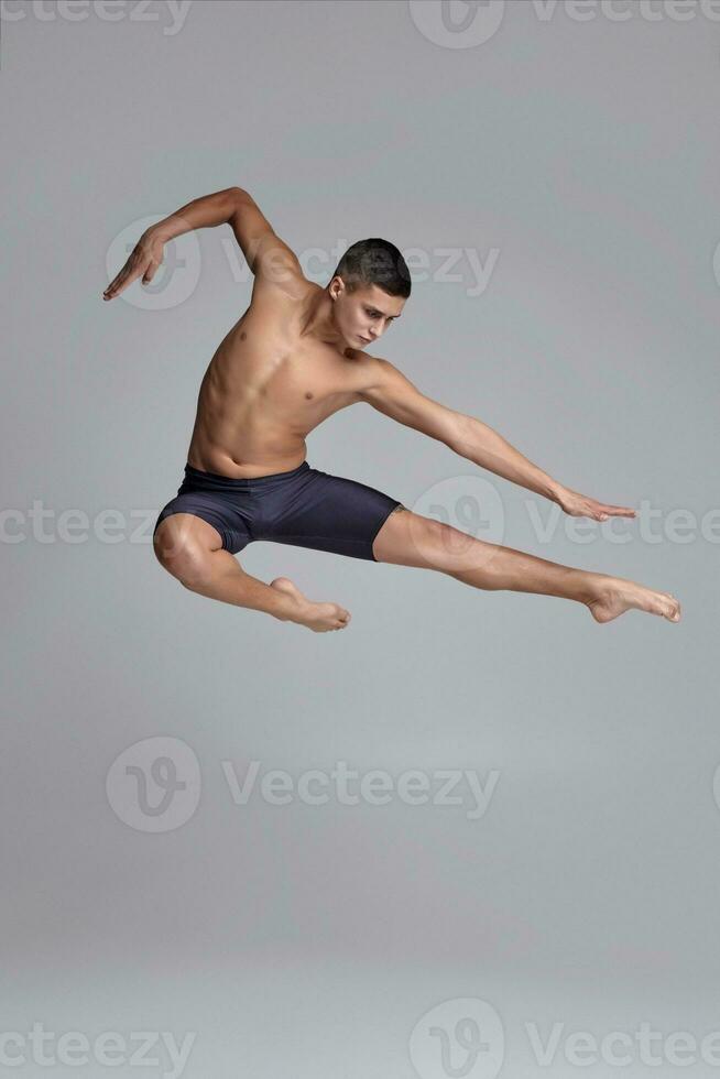 Photo of a handsome man ballet dancer, dressed in a black shorts, making a dance element against a gray background in studio.