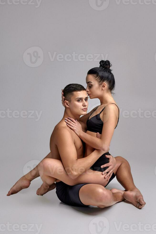 The couple of a young modern ballet dancers in black suits are posing over a gray studio background. photo