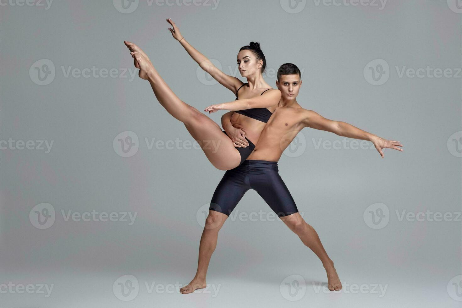 The couple of a young modern ballet dancers in black suits are posing over a gray studio background. photo