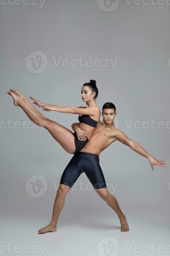 The couple of a young modern ballet dancers in black suits are posing over a gray studio background. photo