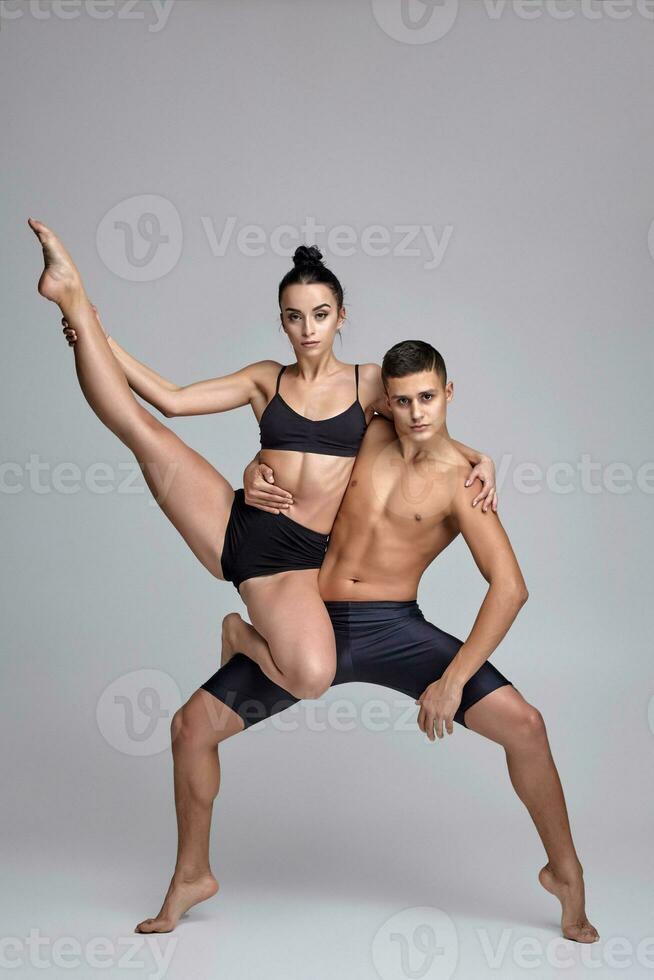 The couple of a young modern ballet dancers in black suits are posing over a gray studio background. photo