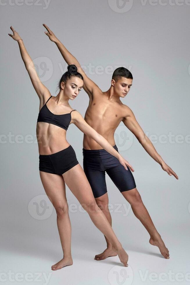 The couple of a young modern ballet dancers in black suits are posing over a gray studio background. photo