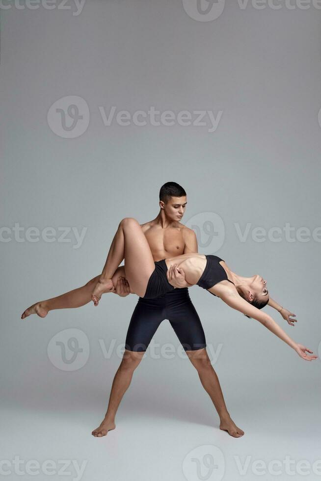 The couple of a young modern ballet dancers in black suits are posing over a gray studio background. photo