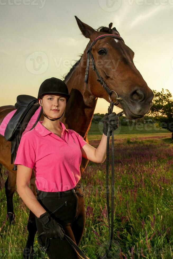 hermosa sonriente niña jockey estar siguiente a su marrón caballo vistiendo especial uniforme en un cielo y verde campo antecedentes en un puesta de sol. foto