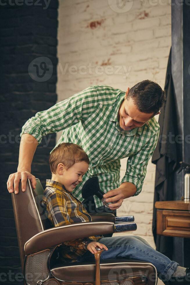 stylish little kid sitting on chair at barbershop with his young father on background photo