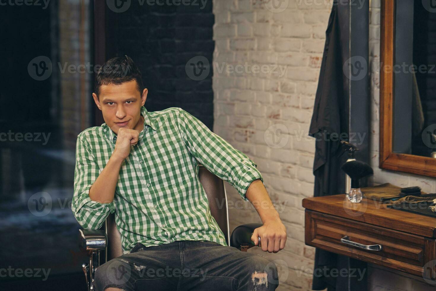 hermoso joven hombre mirando a el cámara y sentado en un silla en a el barbería foto