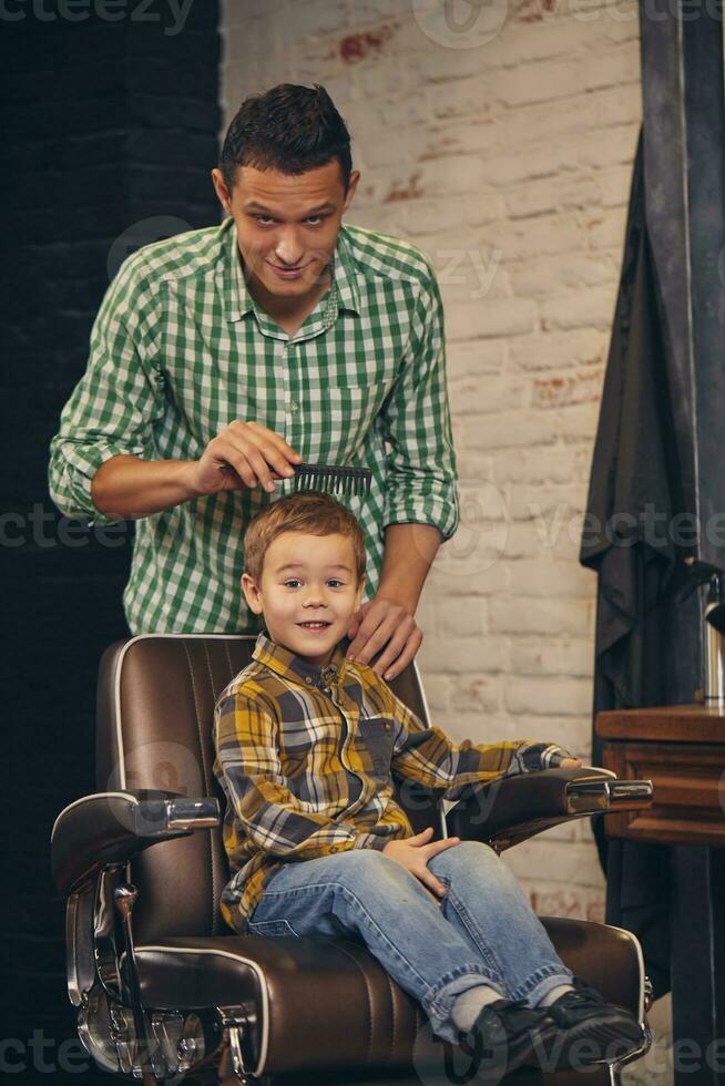 stylish little kid sitting on chair at barbershop with his young father on background photo