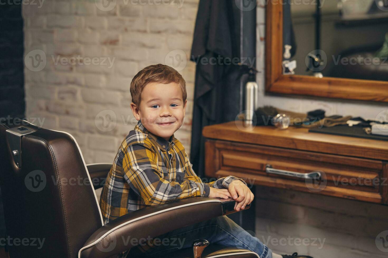 Portrait of a stylish little boy dressed in shirt and jeans in the barbershop, sitting in a chair against the barber's workplace photo