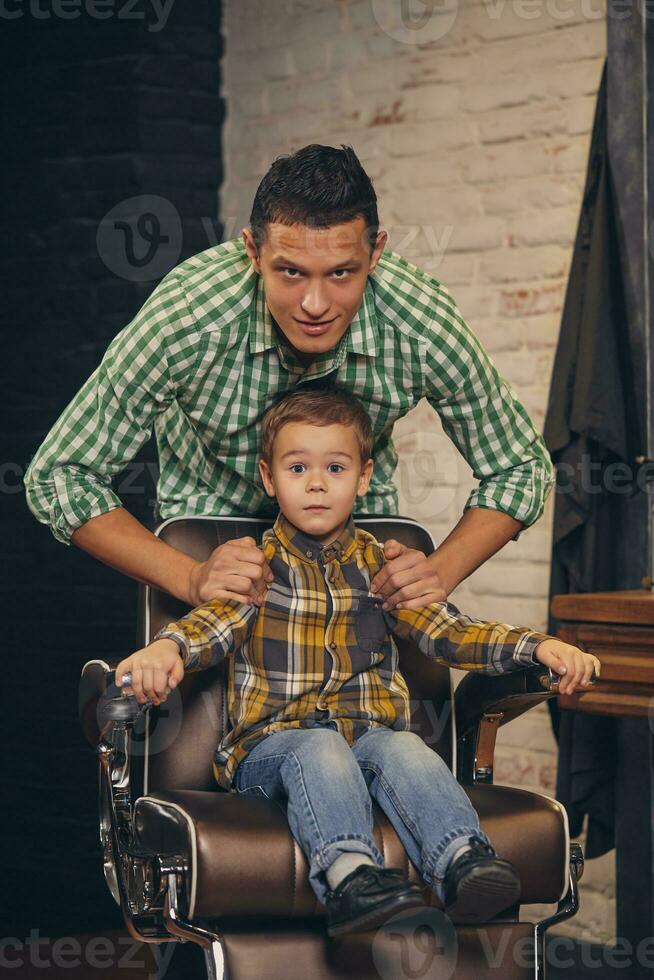 stylish little kid sitting on chair at barbershop with his young father on background photo