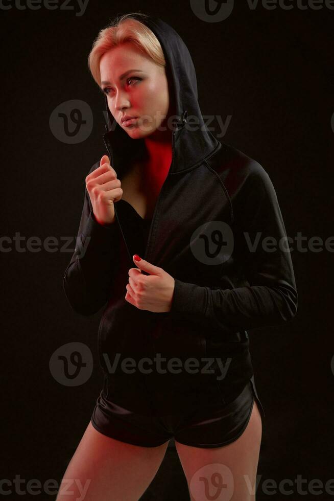 Beautiful seductive sporty girl with the slim sexy figure in the black hoodie, sport underwear and little black shorts is posing in the studio photo