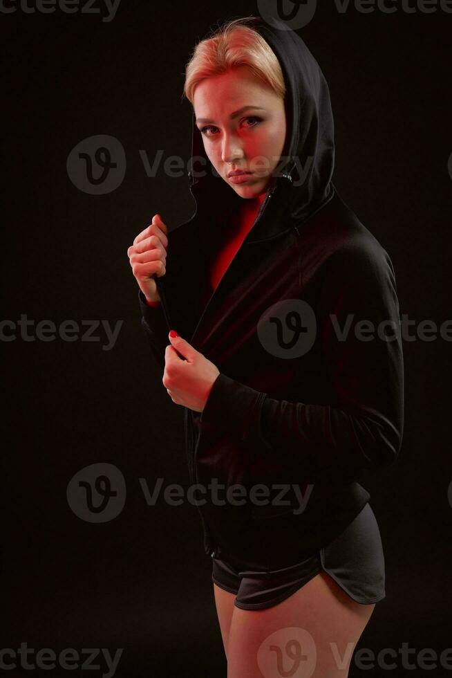 Beautiful seductive sporty girl with the slim sexy figure in the black hoodie, sport underwear and little black shorts is posing in the studio photo