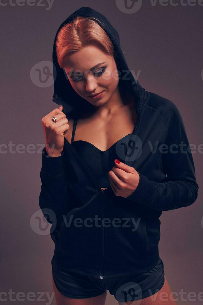 Beautiful seductive sporty girl with the slim sexy figure in the black hoodie, sport underwear and little black shorts is posing in the studio photo