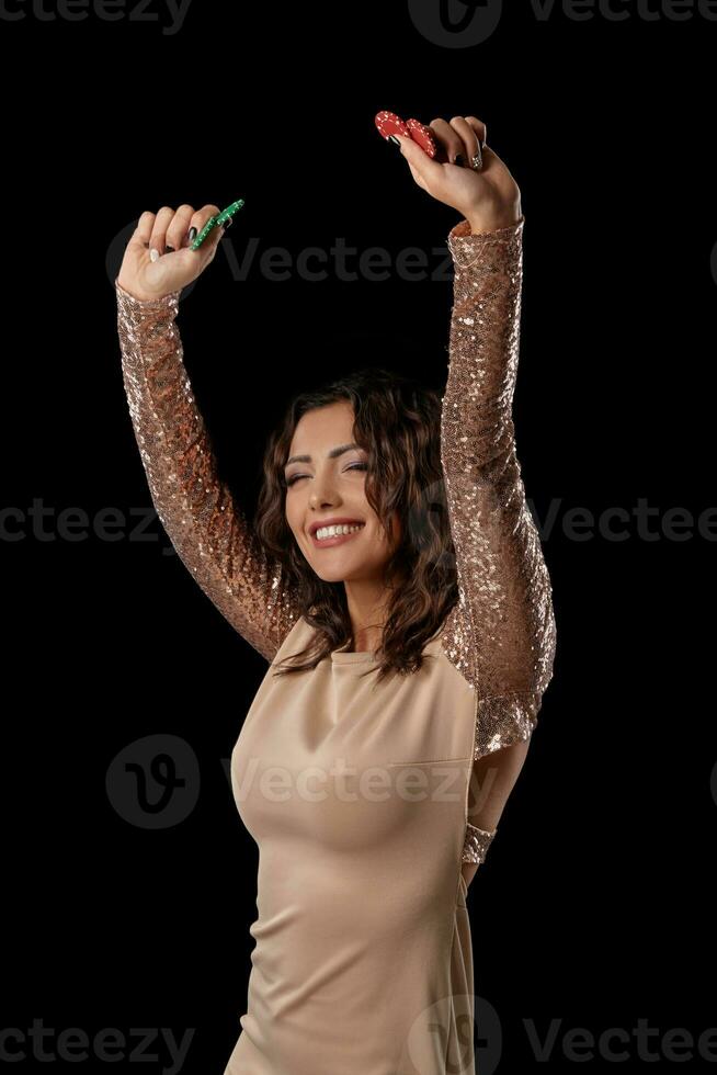 Brunette girl wearing shiny dress posing holding some chips in her hands standing against black studio background. Casino, poker. Close-up. photo