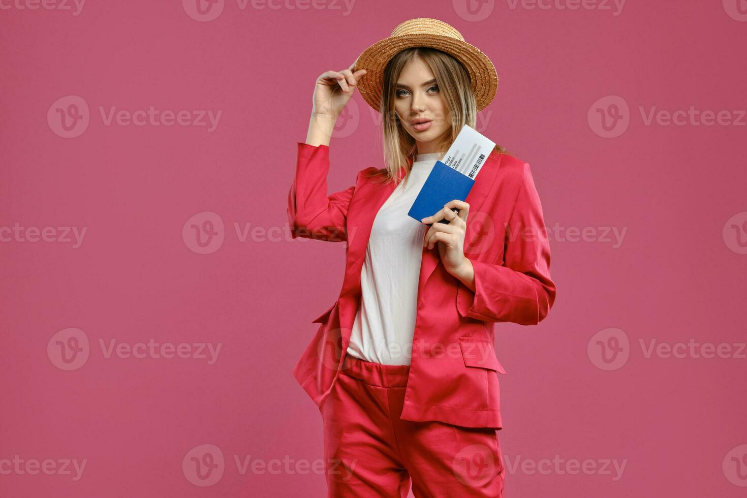 Blonde girl in straw hat, white blouse and red pantsuit. She is holding passport and ticket while posing against pink studio background. Close-up photo