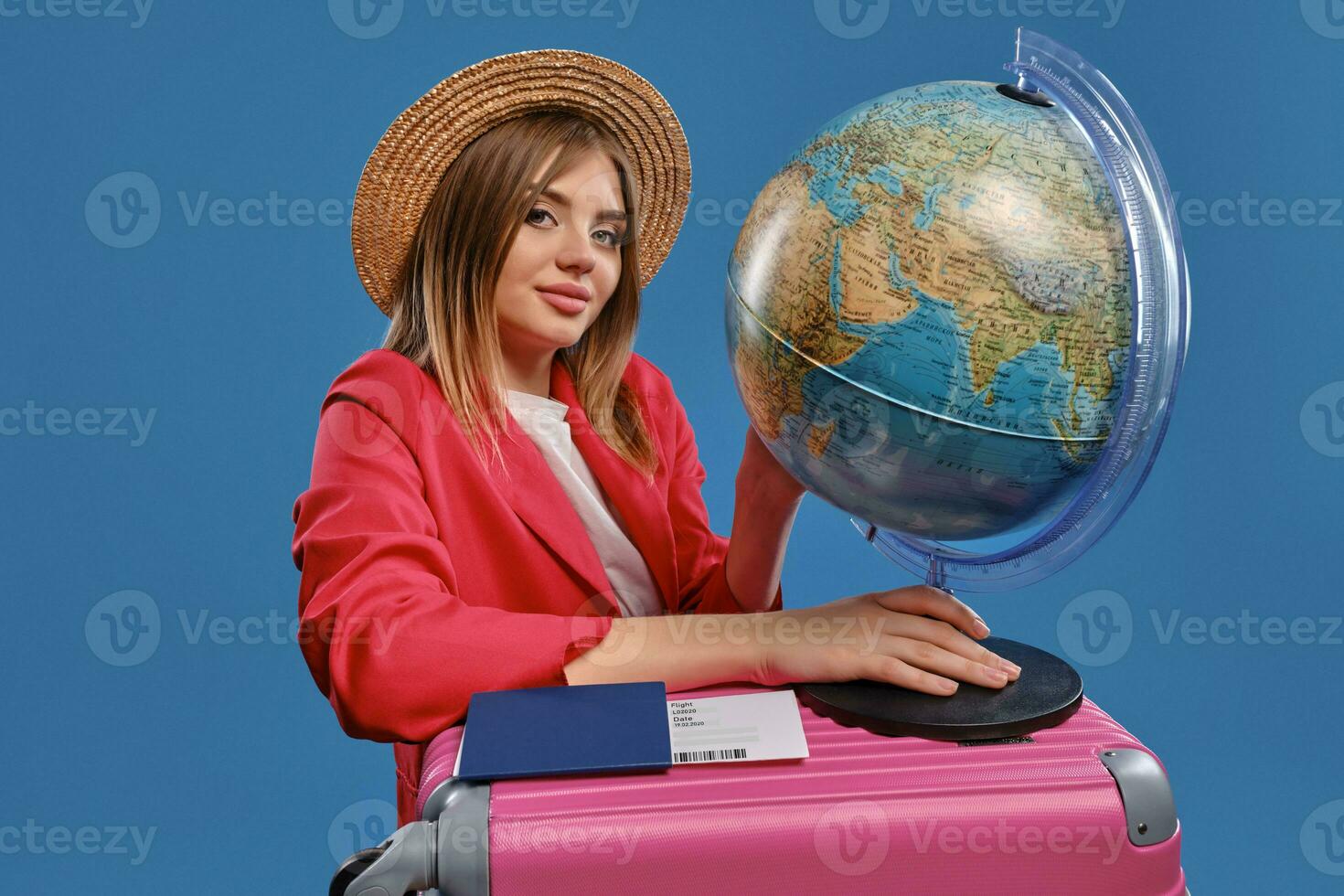 Blonde girl in straw hat, white blouse, red jacket. Holding globe standing on pink suitcase, passport and ticket nearby, posing on blue background photo