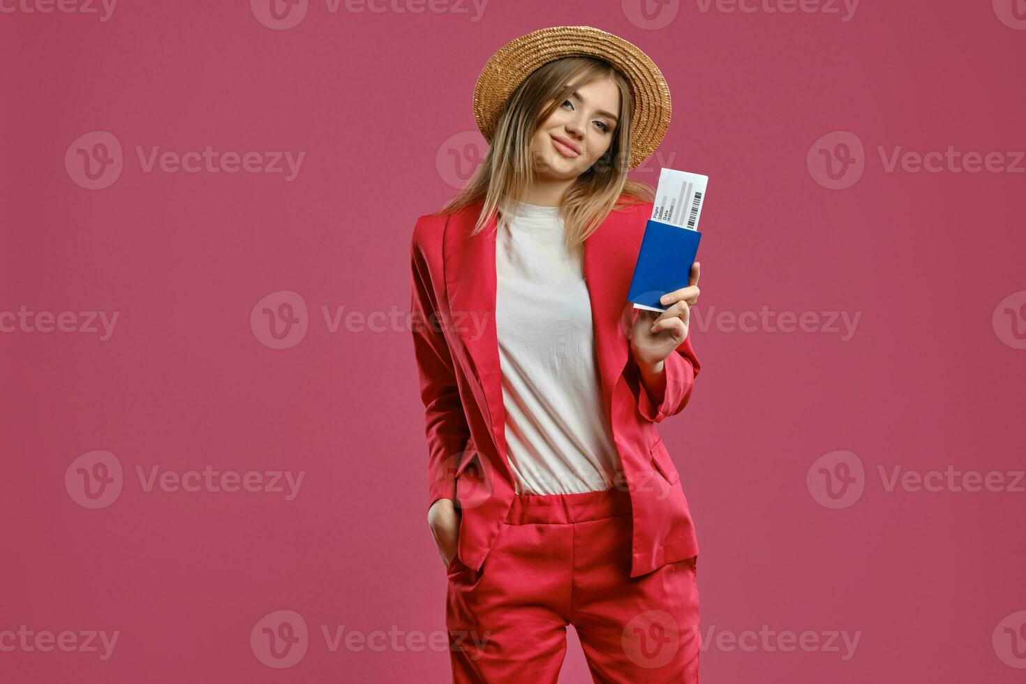 Blonde model in straw hat, white blouse and red pantsuit. She is holding passport and ticket while posing against pink studio background. Close-up photo