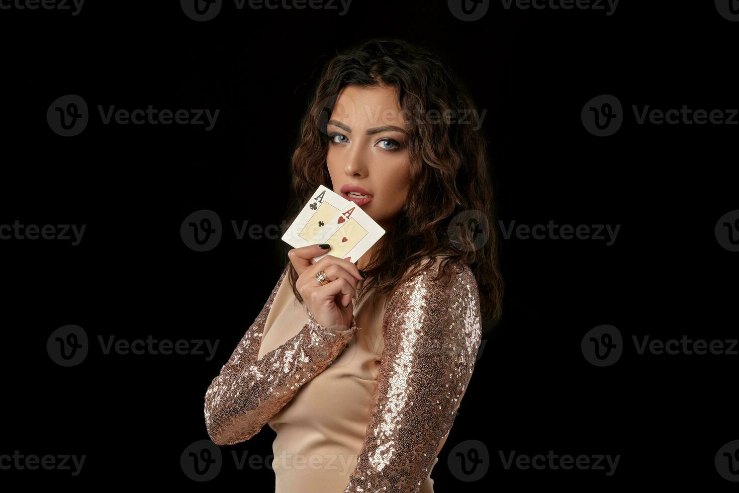 Brunette girl wearing shiny dress posing holding two playing cards in her hand standing against black studio background. Casino, poker. Close-up. photo