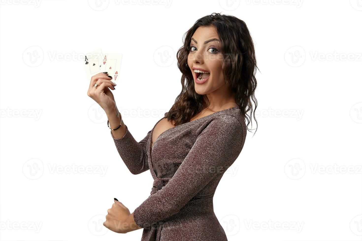 Brunette girl wearing shiny brown dress posing with two playing cards in her hand standing isolated on white background. Casino, poker. Close-up. photo