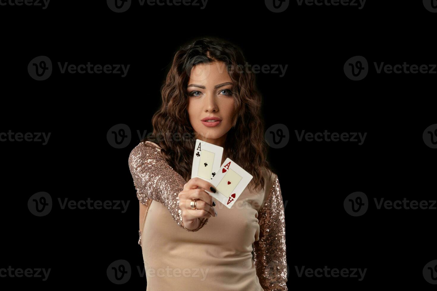 Brunette girl wearing shiny dress posing holding two playing cards in her hand standing against black studio background. Casino, poker. Close-up. photo