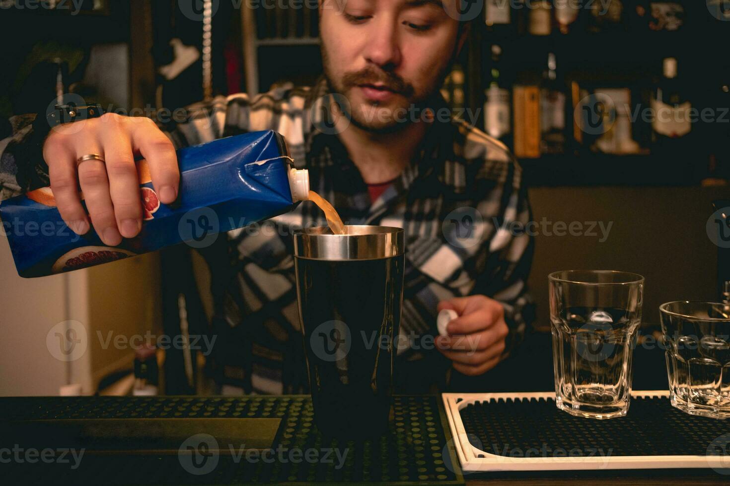 Bearded bartender pouring orange juice from carton to shaker, preparing cocktail photo