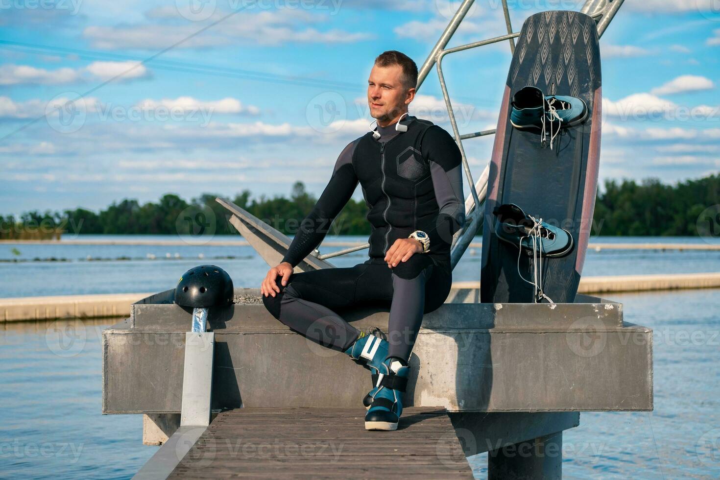 Happy confident man in wetsuit sitting on pier with wakeboard photo