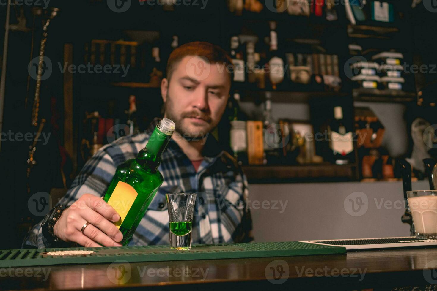 Confident bearded bartender pouring green liquor from bottle into shot glass photo