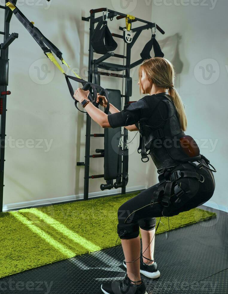 Beautiful blonde woman wearing in sensors and belts, fastens with velcro is practicing EMS fitness in a gym performing traction exercises. photo