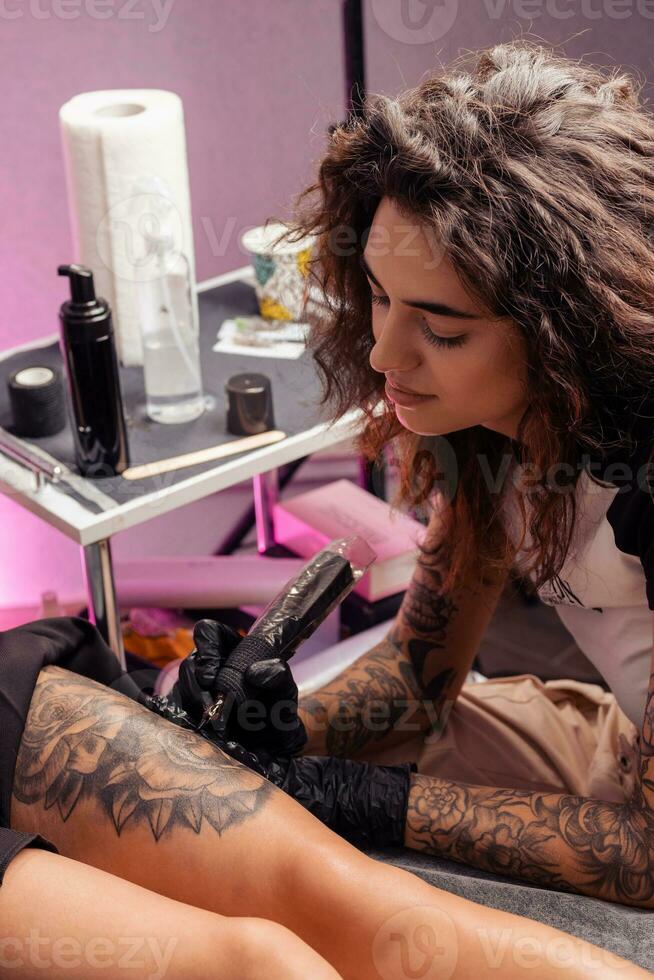 Female tattoo artist using machine to create flower design on client thigh photo