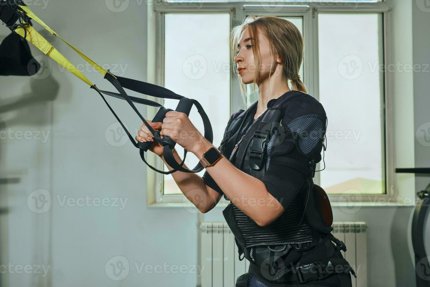 Beautiful blonde woman wearing in sensors and belts, fastens with velcro is practicing EMS fitness in a gym performing traction exercises. photo