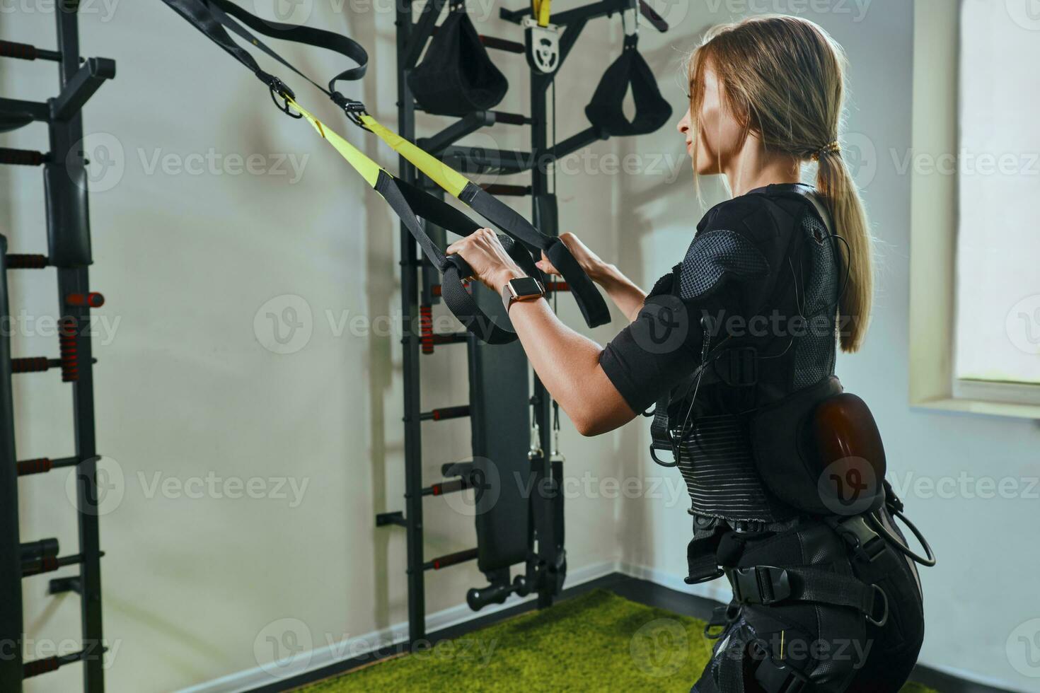 Beautiful blonde woman wearing in sensors and belts, fastens with velcro is practicing EMS fitness in a gym performing traction exercises. photo