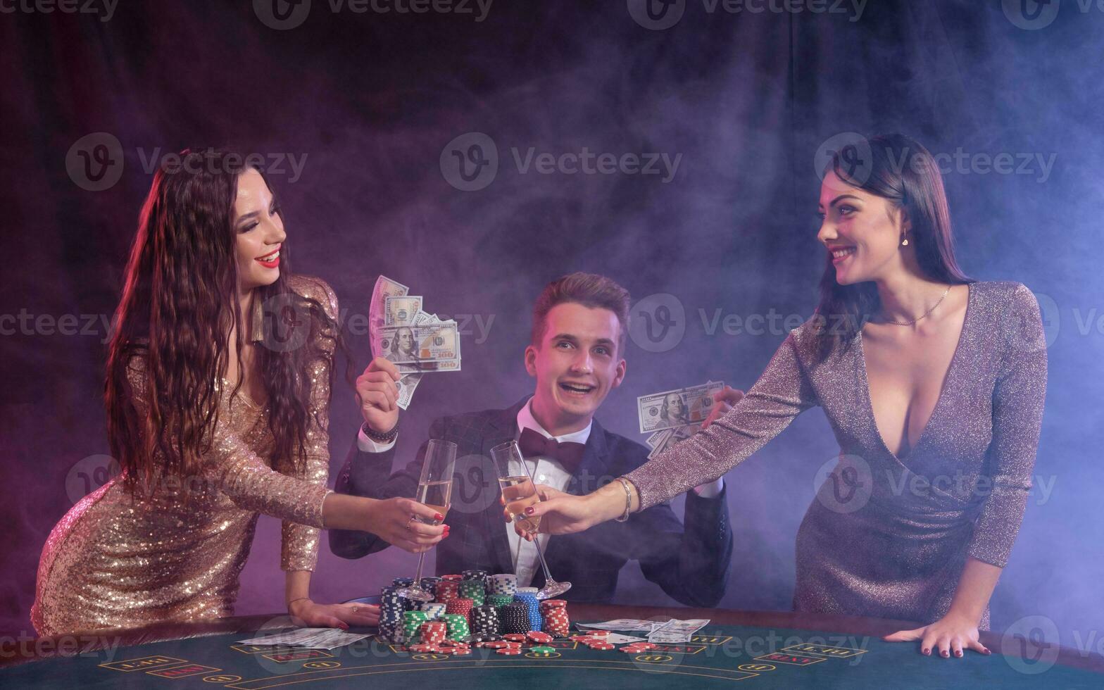 Man playing poker at casino sitting at table with stacks of chips, money, cards. Celebrating win with two women. Black, smoke background. Close-up. photo