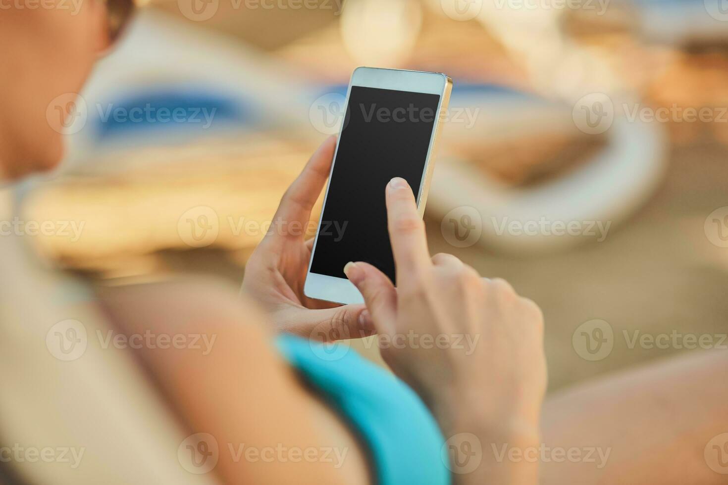 Close up view of a young attractive woman on holiday laying down on a white chaise-longue, holding and using a smartphone. photo