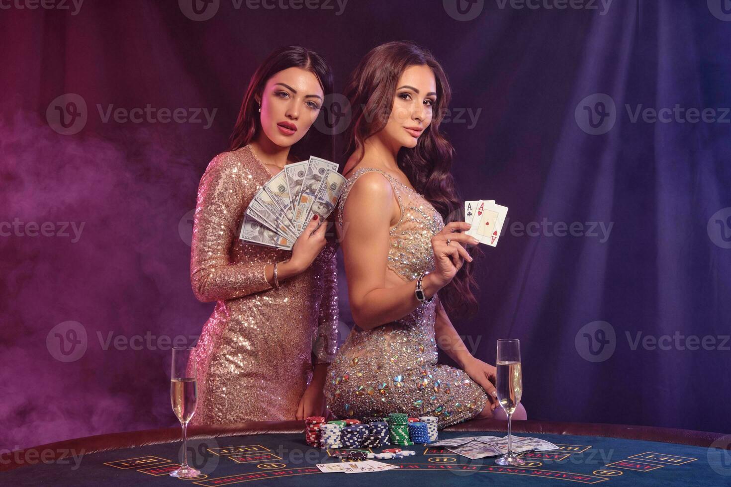 Two women showing cards and money, posing at playing table in casino. Black, smoke background with colorful backlights. Gambling, poker. Close-up. photo