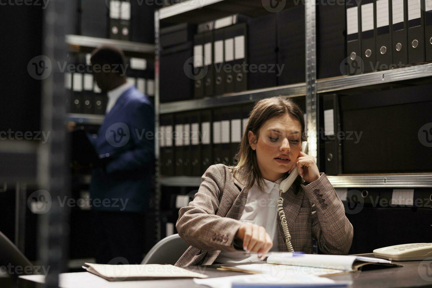 Police officer using landline phone to talk with remote police officer, discussing crime scene evidence in arhive room. Private detective working overtime at criminology report, trying to solve case photo