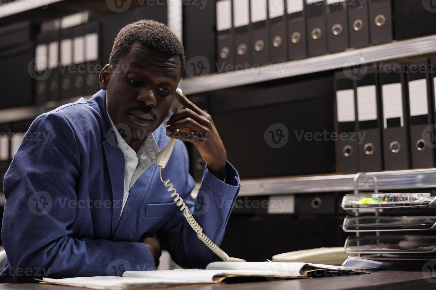 Police officer explaining confidential case to remote inspector using landline phone, working overtime in arhive room. Private investigator checking crime scene evidence. Investigation concept photo