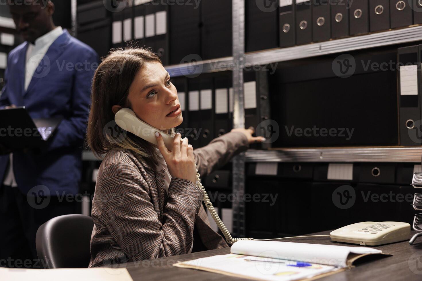 Inspector explaining police report using landline phone, working overtime at criminal investigation in arhive room. Private detective analyzing crime scene evidence searching for new clues photo