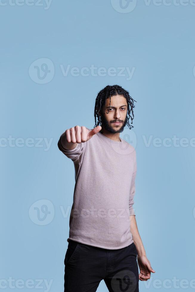 Cool arab man showing neutral thumb sign with hand and looking at camera. Young person raising eyebrow and making satisfaction normal level gesture with finger on blue background photo