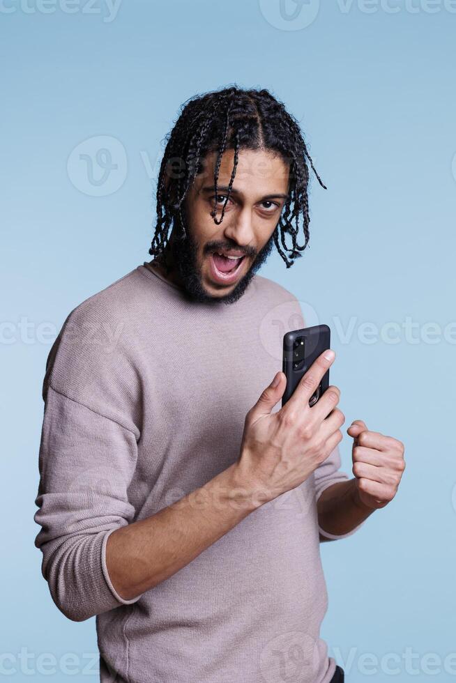 Excited arab man checking message on smartphone and showing winner gesture with clenched fist portrait. Person using mobile phone and looking at camera with happy expression photo