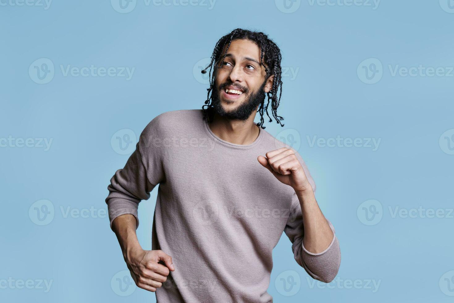 Cheerful arab man dancing with happy facial expression and looking away with smile. Young adult arabian handsome person in good mood making moves with hands on blue background photo