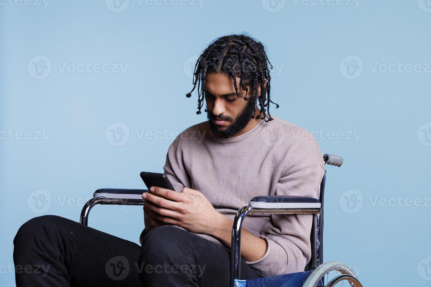 Arab man sitting in wheelchair while scrolling social media on smartphone. Young person with physical impairment sending online message and browsing internet on mobile phone photo